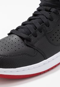 Zapatilla alta de cuero negro con perforaciones, cordones negros planos, mediasuela blanca en contraste y suela de goma roja.