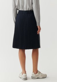 Jupe plissée bleu marine avec une taille ajustée, en tissu texturé, portée avec un pull gris et des baskets de couleur claire.