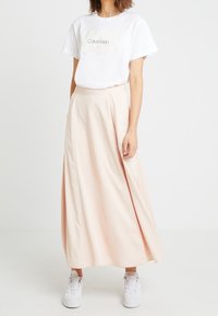 Světle růžová maxi sukně s hladkou texturou, spárovaná s bílým tričkem Calvin Klein. Modelka v bílých teniskách.