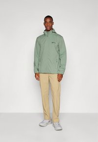 Casaco verde claro com fecho éclair e capuz, com um logótipo no peito; usado com calças bege e ténis cinzentos. Design simples e funcional.