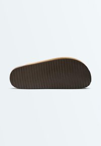 Semelle plate marron d'une chaussure sans lacets avec des rainures horizontales pour l'adhérence, présentée sur un fond clair uni.