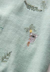 Puuvillane kangas helemintrohelises toonis, millel on tikitud mustrid, sealhulgas puuoksal rippuv koala ja dekoratiivsed rohelised lehed.