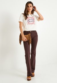 Camiseta de algodón claro con estampado gráfico en rosa y azul, combinada con pantalones acampanados marrón oscuro y un clutch de ante marrón con flecos.