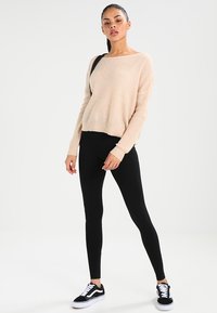 Pull en tricot beige à coupe décontractée, associé à un legging noir et des baskets noires. La texture est douce, avec des manches longues et un large col.