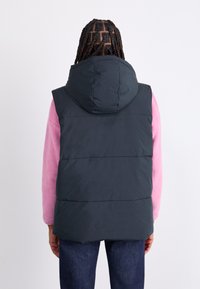 Gilet imbottito blu navy con cappuccio, caratterizzato da un design trapuntato e cuciture anteriori, indossato sopra un top a maniche lunghe in pile rosa.