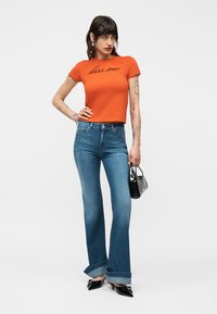 Γυναίκα που φοράει πορτοκαλί μπλουζάκι με την επιγραφή "φίλα με", τζιν ψηλόμεσο με φαρδιά μανσέτα, μαύρες μυτερές γόβες, κρατώντας μικρή μαύρη τσάντα.