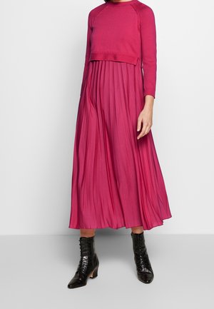 Vrouw die een felroze gebreide top met lange mouwen draagt en een bijpassende geplooide midirok, gecombineerd met zwarte veterlaarzen tot aan de enkel.