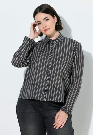 Woman with short dark hair wearing a black and white vertical striped button-up shirt and dark pants, posing against a white background.