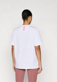 Bijela pamučna majica opuštenog kroja, kratkih rukava, s ružičastom etiketom na stražnjem dijelu vrata. Glatka tekstura, bez vidljivih uzoraka ili naglasaka.