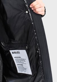 Čierna izolačná bunda zvnútra s prešívanou štruktúrou, so zacvakávacími uzávermi a štítkom s názvom značky a pokynmi k starostlivosti.