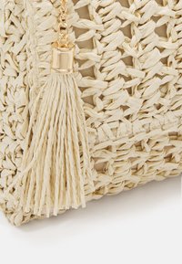 Beżowa torba pleciona ze słomy o fakturowanej powierzchni; posiada długi, zwisający chwost oraz złocony okrągły detal metalowy.
