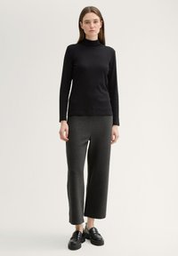 Melna ilga piedurkne turtlenecka augšdaļa, kombinēta ar tumši pelēkiem platām biksēm. Modelis valkā melnas platformas kurpes. Gluda tekstūra, pieguļošs dizains.