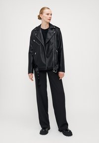 Svart skinnbikerjakke med sølvglidel�åser, sammen med svarte pinstripe vide bukser og kraftige svarte sko.