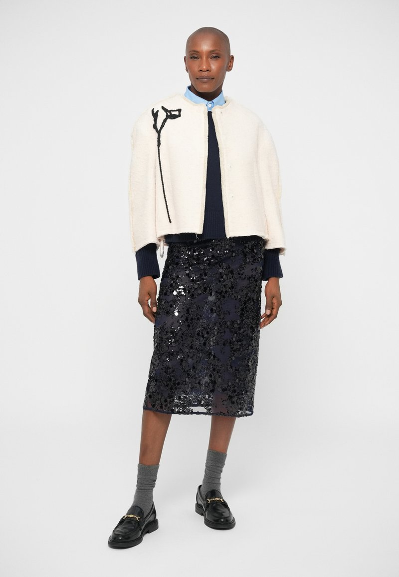 Cappotto in misto lana bianco con ricami floreali neri, abbinato a una gonna a tubino blu navy in paillettes. Il modello indossa calze grigie e mocassini neri.