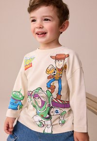 Svetle béžové tričko s dlouhým rukávem, které na přední straně zdobí barevné potisky postav z Toy Story, jako jsou Woody, Buzz Rakeťák a jedna postava z mimozemské kultury.