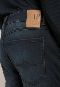 Jeans de mezclilla azul oscuro con una etiqueta de cuero que lleva "JP 1880". El diseño incluye un bolsillo trasero con costuras curvadas y hilo de contraste.