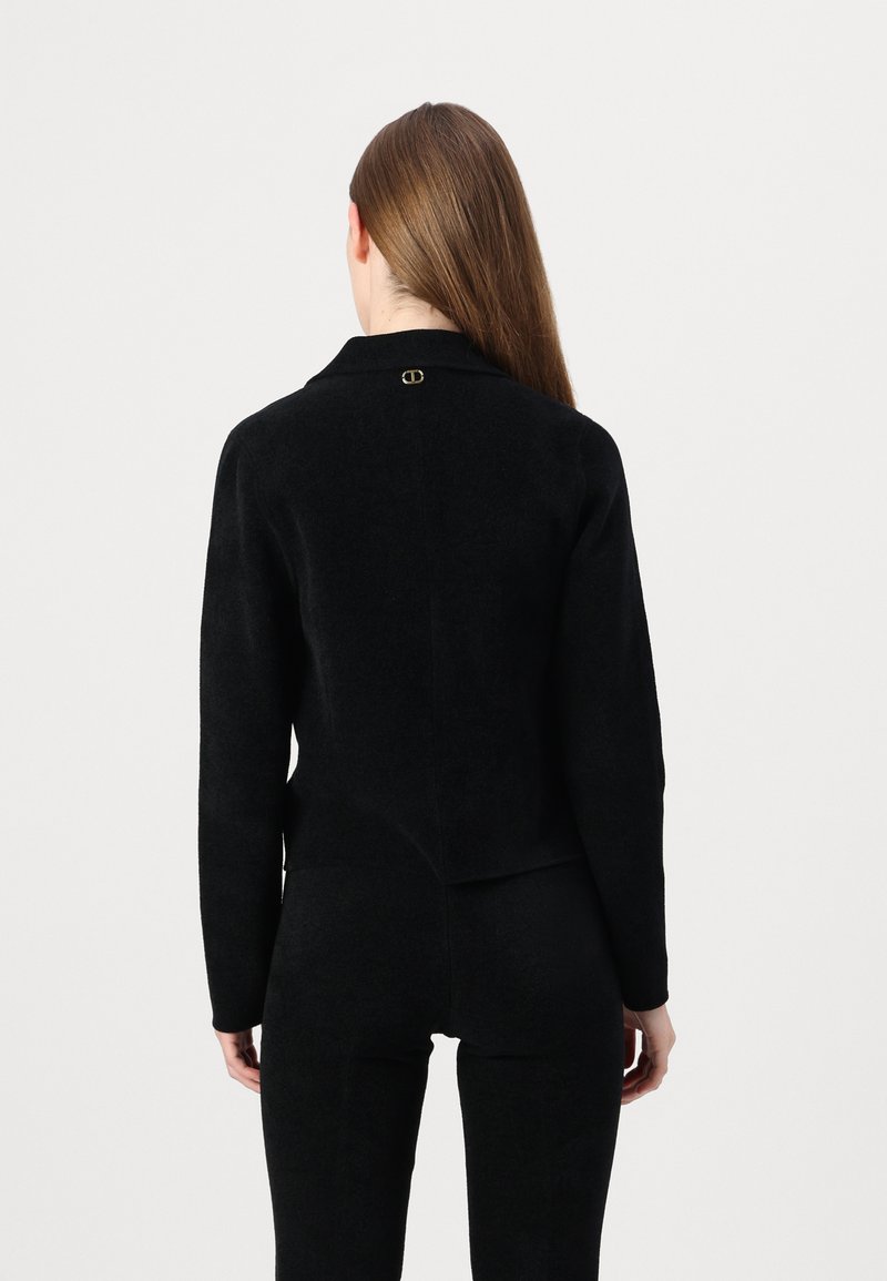 Schwarze Wollmischung Jacke mit Kragen, tailliertem Design, Rückenschlitz und goldfarbenem Logodetail am Hals. Weiche Textur und strukturierte Silhouette.