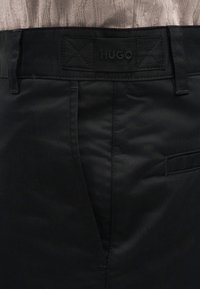 Κοντινό πλάνο της μέσης μαύρων παντελονιών με θηλιές για ζώνη, ετικέτα με το εμπορικό σήμα "HUGO" και μέρος μπεζ πλεκτού πουλόβερ μισό σφιγμένο μέσα.