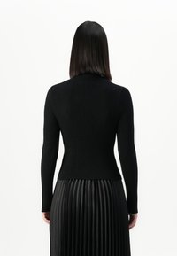 Woman with straight dark hair wearing a black ribbed long-sleeve top and a black pleated skirt, shown from the back against a white background.