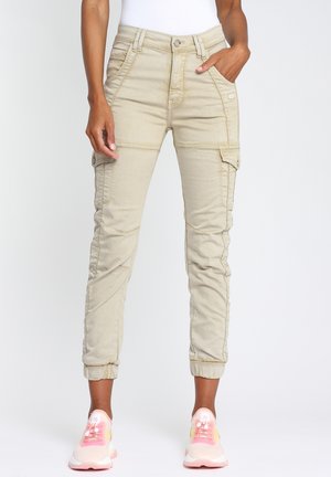 Pantalon cargo beige avec des poignets élastiques porté par une personne ayant une main dans la poche et des baskets roses et blanches sur un fond blanc uni.