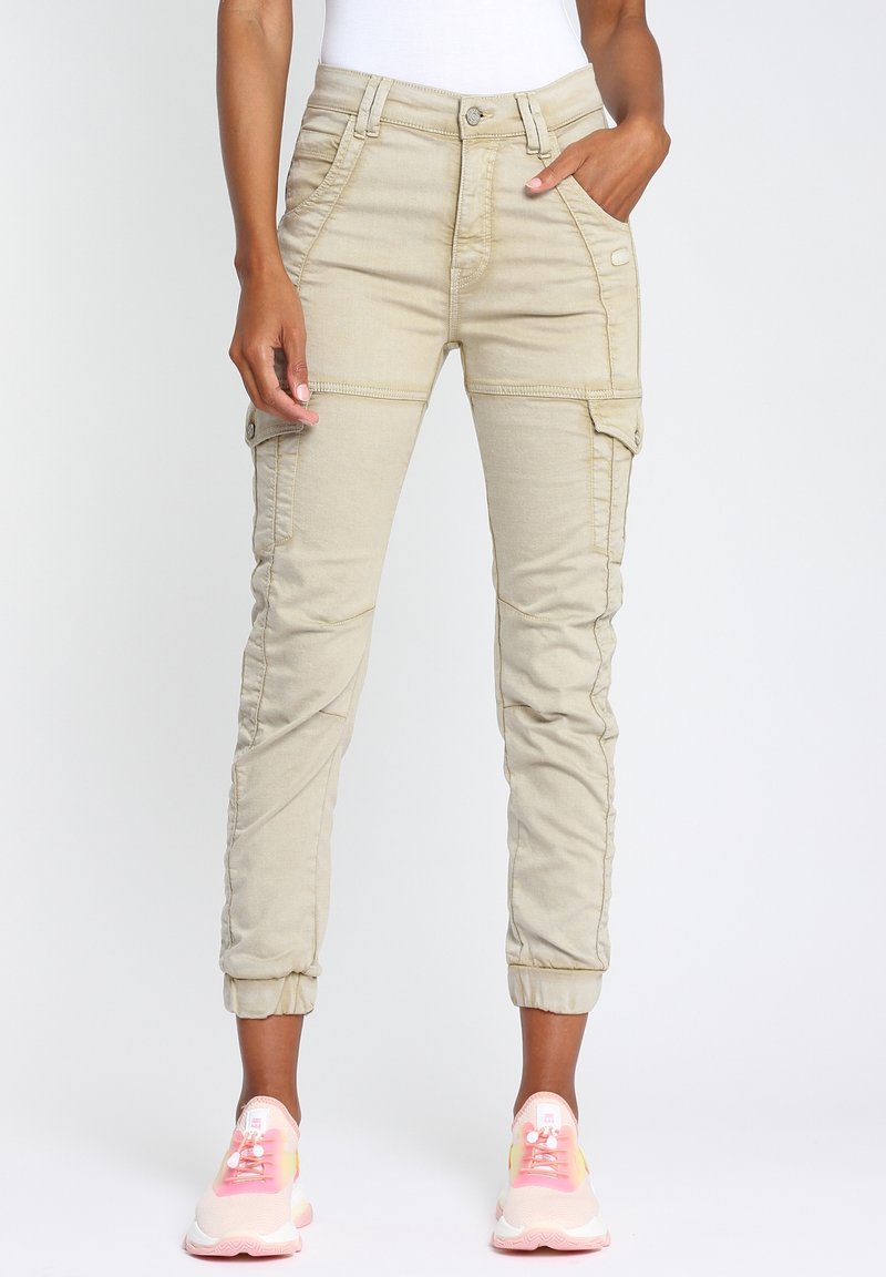 Pantalon cargo beige avec des poignets élastiques porté par une personne ayant une main dans la poche et des baskets roses et blanches sur un fond blanc uni.