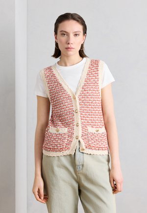 Kvinde iført en knappet, tekstureret strikket vest med røde og cremefarvede striber over en hvid t-shirt og lyse beige bukser, stående op ad en grå væg.