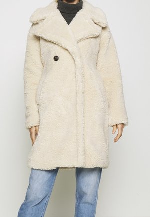 Personne portant un manteau en polaire texturée crème, par-dessus un col roulé foncé et un jean bleu, sur un fond clair uni.