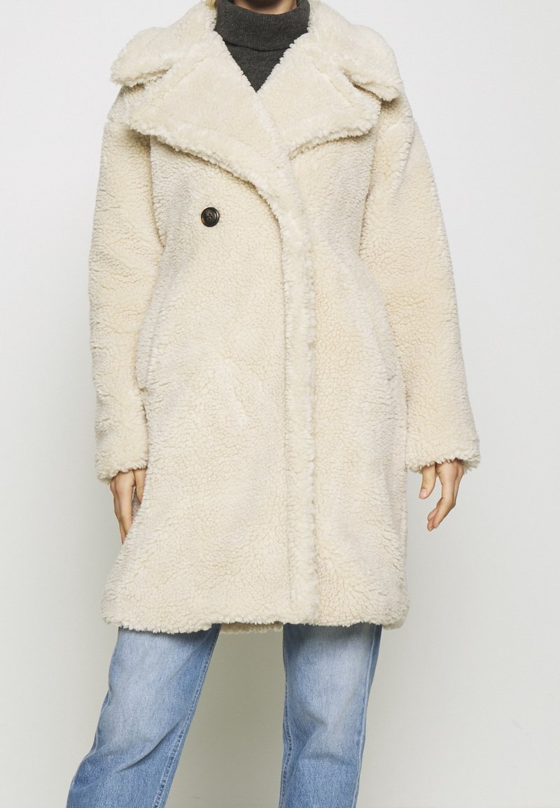 Personne portant un manteau en polaire texturée crème, par-dessus un col roulé foncé et un jean bleu, sur un fond clair uni.