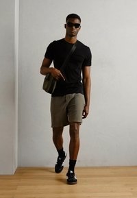 Sort T-shirt, olivenfarvede shorts og sorte sneakers med hvide detaljer; modellen bærer solbriller og har en lille crossover-taske.