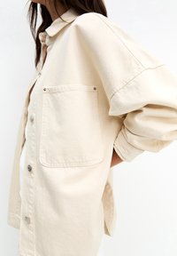 Beige katoenen shirt met een kraag, knoopsluiting en een groot borstzakje. Heeft oversized mouwen en een ontspannen pasvorm.