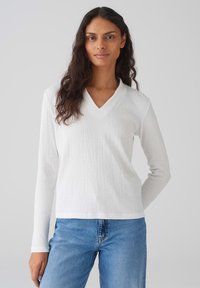 Weißes, langärmeliges V-Ausschnitt-Oberteil aus strukturiertem Stoff, kombiniert mit blauen Jeans. Das Oberteil hat einen lässigen Schnitt und dezente Nahtdetails.