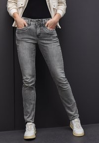 Jeans skinny de mezclilla gris con una cintura de media altura, bolsillos frontales y cierre de botón. Llevados con zapatillas deportivas blancas.