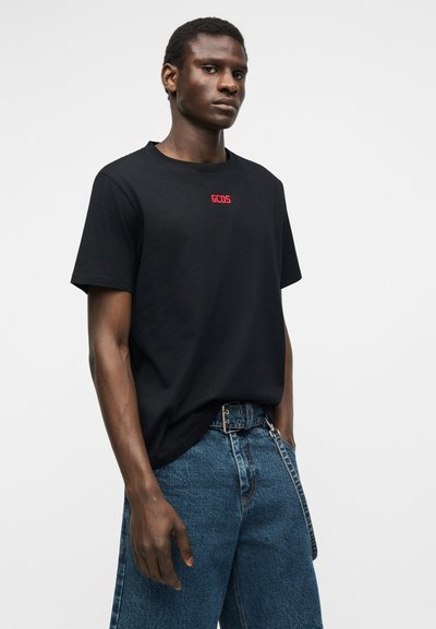 Νέος άνδρας που φοράει μαύρο t-shirt GCDS και μπλε τζιν με μαύρη ζώνη και αλυσίδα ως αξεσουάρ, στέκεται μπροστά σε ένα απλό λευκό φόντο.