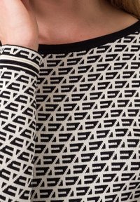 Schwarz-weiß gemusterter Pullover mit geometrischem Design auf einem strukturierten Strickstoff. Der Ausschnitt und die Bündchen haben kontrastierende schwarze Akzente.