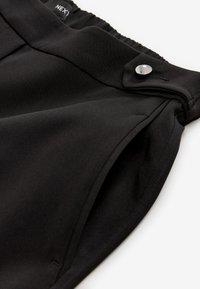 Pantalon noir taille haute avec ceinture élastique, poche latérale et détail bouton-pression argenté sur la languette de la ceinture.