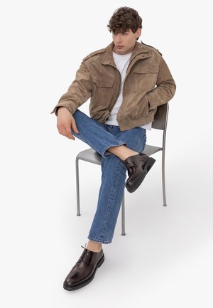 Mężczyzna siedzi na krześle ze skrzyżowanymi nogami, ubrany w brązową zamszową kurtkę, białą koszulkę, niebieskie dżinsy i ciemnobrązowe skórzane buty.