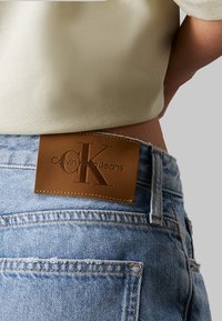 Jeans de mezclilla azul claro con una etiqueta de cuero marrón que muestra "Calvin Klein Jeans". Presenta costuras de contraste y un diseño clásico de cinco bolsillos.