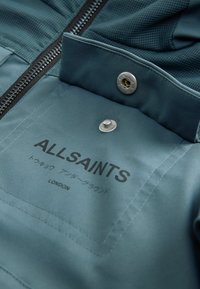 Modra jakna z zadrgo, izdelana iz teksturirane tkanine, s srebrnim okovjem in natisnjenim logotipom "ALLSAINTS" v črni barvi. Preprost dizajn ovratnika.