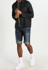 Svart, lätt jacka med en framficka, som bärs över en grön skjorta. Matchad med denimshorts och vita sneakers med uppvikta fållar.