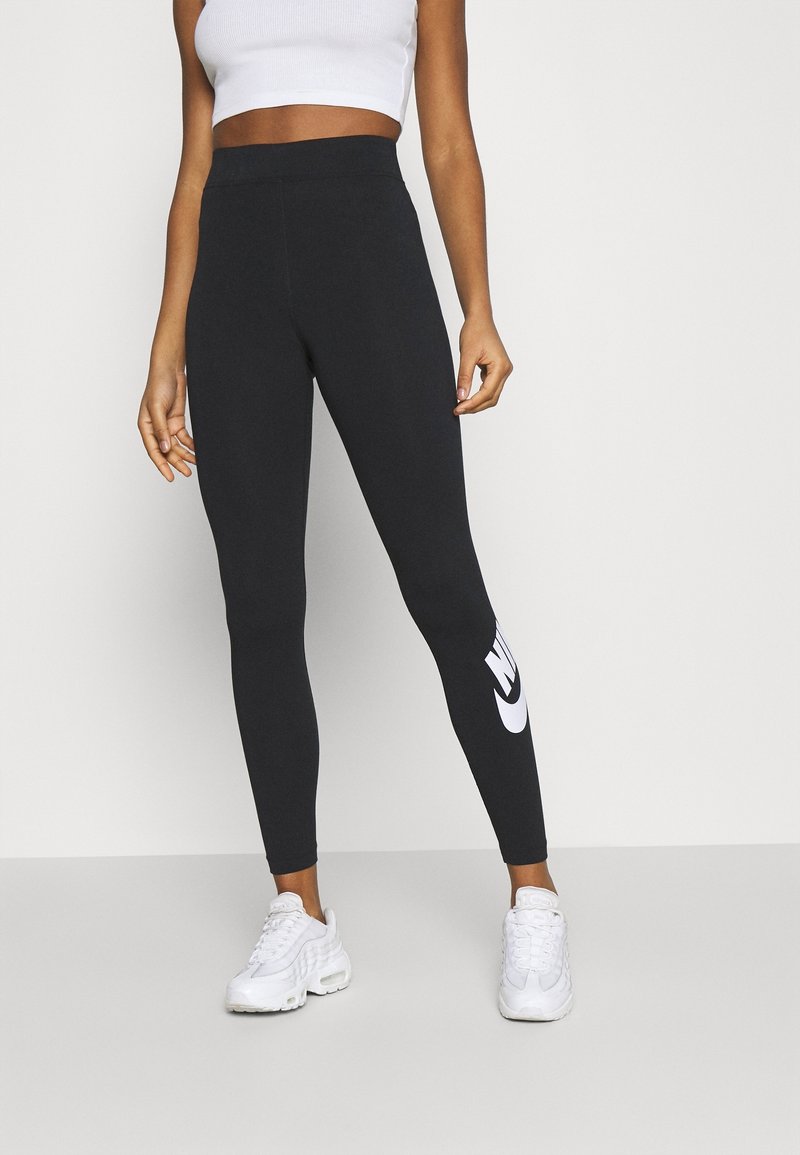 Nainen, jolla on päällään mustat Nike-legginssit, joissa on valkoinen logo säärellä, valkoinen crop-top ja valkoiset lenkkarit, seisoo vaaleaa taustaa vasten.