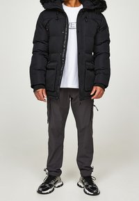 Chaqueta de abrigo negra con capucha con pelo sintético, cierre frontal y múltiples bolsillos, combinada con pantalones cargo oscuros y zapatillas negras y grises.