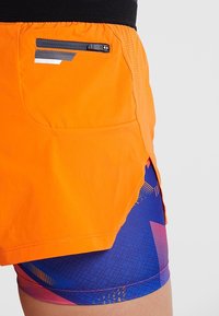 Orange atletiska shorts med en blixtlåsficka och ett blått mönstrat innerfoder med geometriska mönster och kontrasterande detaljer.