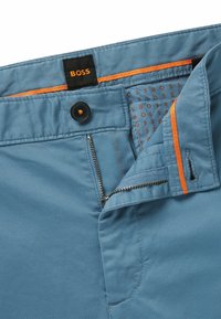 Pantalon bleu clair avec une texture lisse. Présente une étiquette noire avec "BOSS", des accents orange, une fermeture éclair et un bouton.