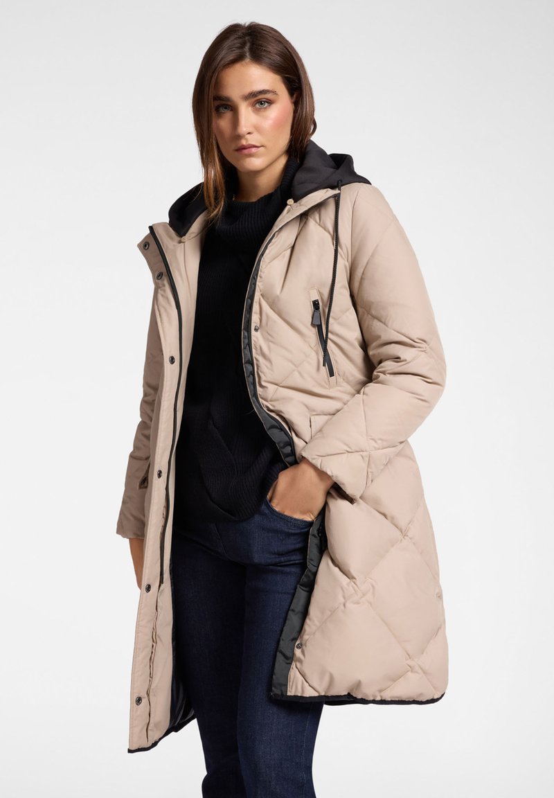 Béžový prošívaný dlouhý kabát s černou kapucí, s kapsou na zip a zapínáním na druky, nošený přes tmavý svetr a džíny.