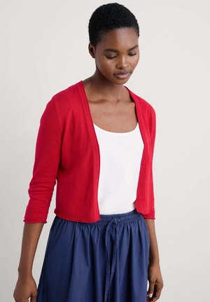 Gilet rouge en tricot avec des manches trois-quarts, porté sur un haut blanc. Assorti à une jupe bleu marine avec une taille à cordon.