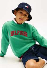 Αγόρι καθισμένο, φοράει πράσινο φούτερ με την επιγραφή "HILFIGER" σε κόκκινο, σκούφο κουβά σε μπλε ναυτικό χρώμα και σκούρα τζιν σορτς, μπροστά σε λευκό φόντο.
