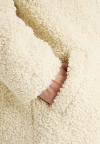 Krämsfärgad fluffig jacka i en texturerad, lockig tyg. Har en ficka på framsidan, synlig söm och en manschett vid ärmen.