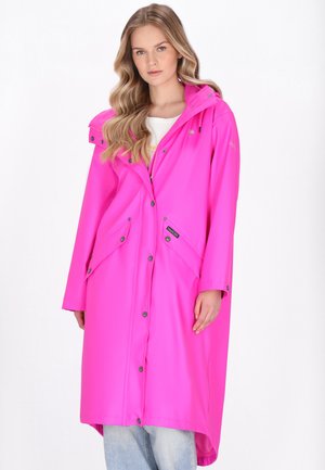 Hosszú szőke hajú nő élénk rózsaszín esőkabátban, patentokkal és ferde zsebekkel, fehér háttér előtt állva.