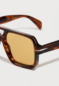 Kvadrātveida saulesbrilles ar dzintara krāsas lēcām un bruņurupuča raksta rāmi, kas atpūšas uz baltas virsmas.