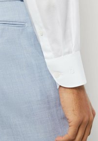 Weißes Hemd mit strukturiertem Muster, geknöpfter Manschette, kombiniert mit hellblauem Strukturdruckstoff, zeigt einen Teil einer Hand, die dagegen lehnt.